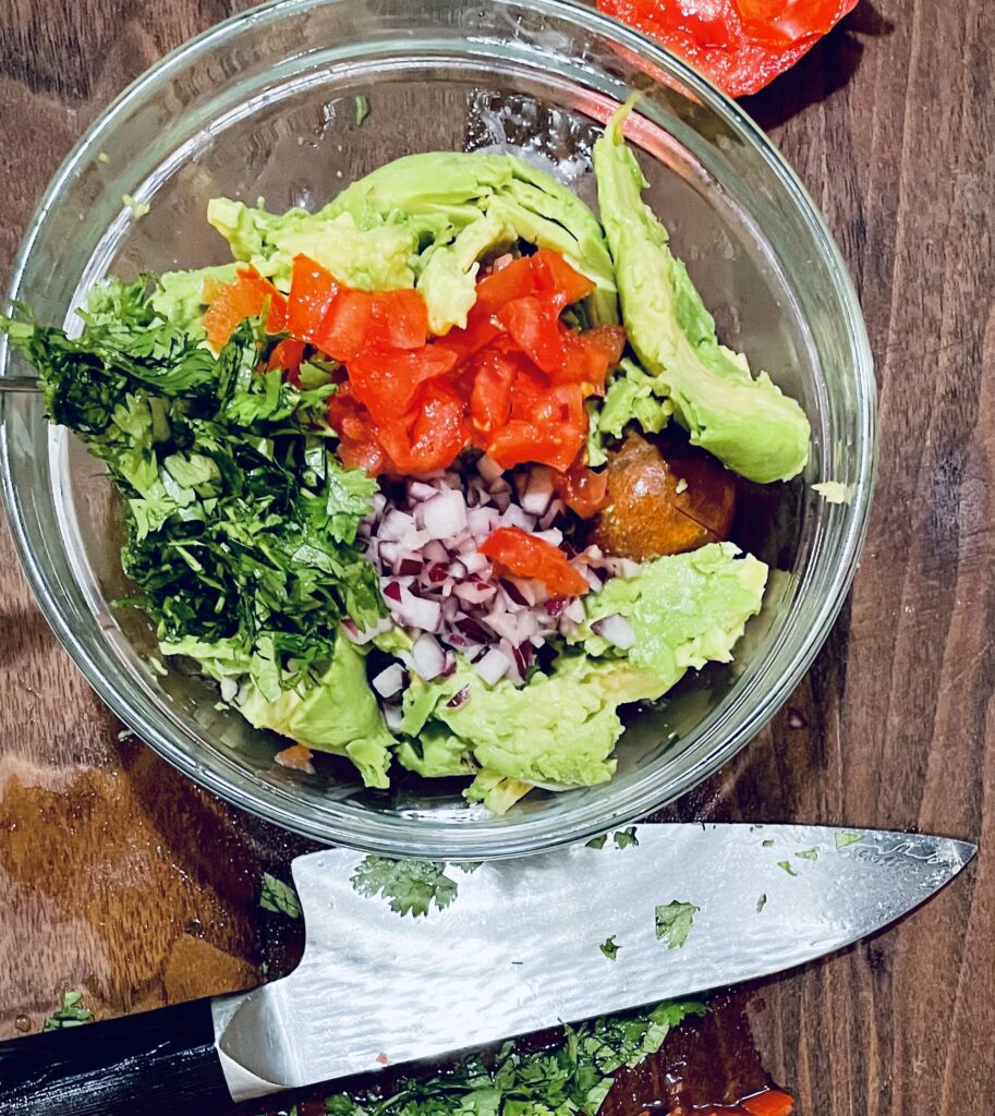 Added chopped cilantro to bowl that has: Red onions, tomatoes and avocados in clear bowl. There is also a knife next to the bowl.
