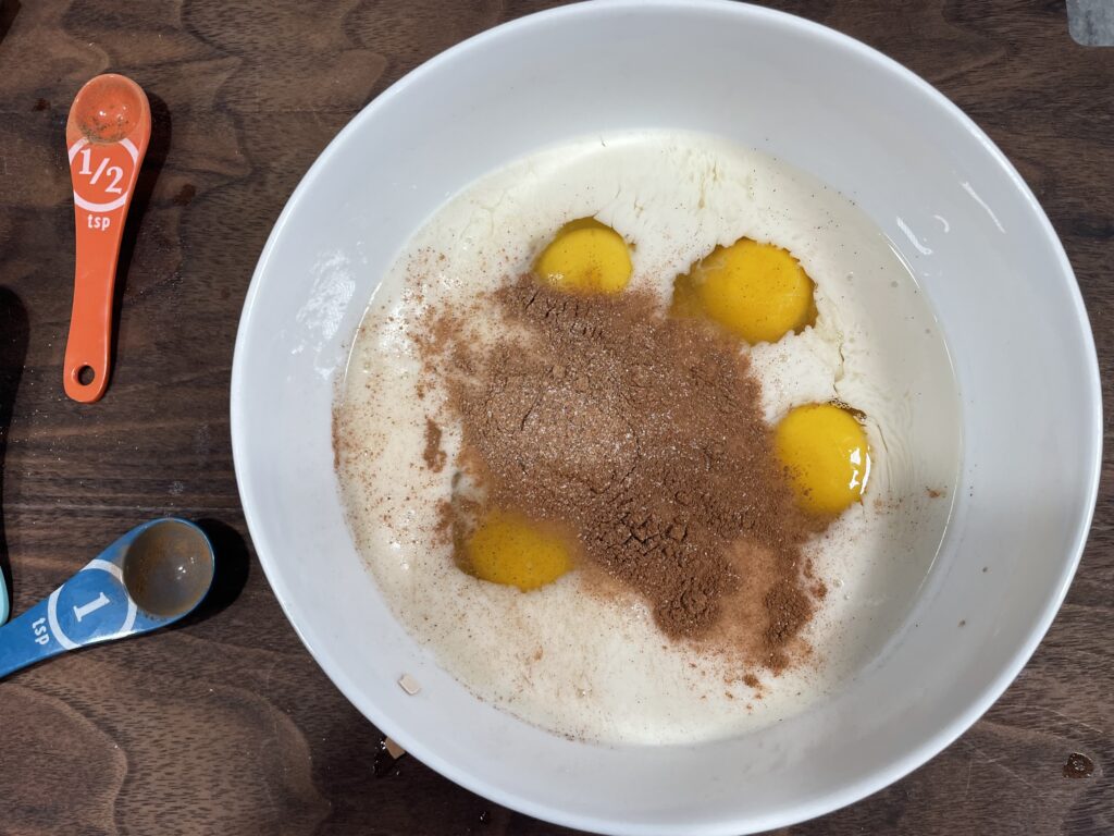 eggs, milk, vanilla, cinnamon, nutmeg and Bailey’s Irish Cream. In a bowl on top of a brown cutting board. 2 measuring spoons next to the bowl.
