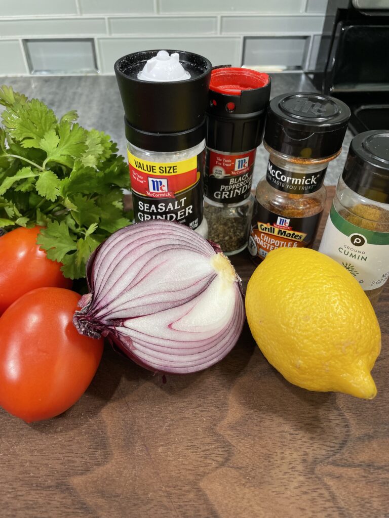 Plum tomatoes or Red Onion Fresh Cilantro Chipotle Ground Cumin Black Pepper Sea Salt Lemon Juice Olive Oil All ingredients are on a brown cutting board