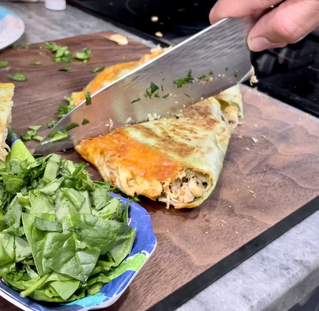 Cutting Quesadilla in brown cutting board. The is some chopped up spinach in a dish next to it. 