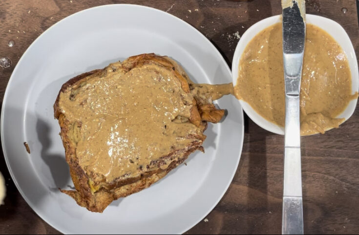 Peanut butter on a small bowl with spreading knife on top of small bowl. 2 slices on french toast covered with peanut butter. On a gray plate.
